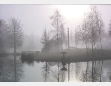 Ein nebliger Park mit einem ruhigen Teich, in dem Bäume und ein Zierbrunnen im Wasser reflektiert werden; im Hintergrund steht eine Statue auf einer hohen Säule neben einer Bank auf einer kleinen Erhebung.