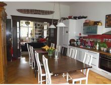 Esszimmer mit integriertem Küchenbereich, einem langen Holztisch mit Stühlen in der Mitte und einer weißen Einbauküche mit roten Fliesen im Hintergrund. Im angrenzenden Raum befinden sich ein Sofa, Regale und helle Fenster, die durch eine dunkle Holzrahmentür sichtbar sind.