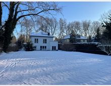 Verschneiter Garten mit großer Rasenfläche vor einem weißen, zweigeschossigen Wohnhaus mit flachem Walmdach und breiten Fenstern. Im Hintergrund stehen weitere Häuser hinter einer Hecke sowie hohe, kahle Bäume unter blauem Himmel.