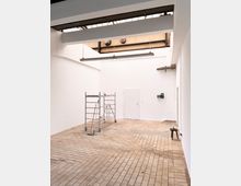 Großer, leerer Raum mit weißen Wänden und einem Boden aus hellbraunen Fliesen. Ein Rollgerüst steht in der Mitte, an der Decke befinden sich freiliegende Träger und eine verglaste Dachkonstruktion, die Tageslicht hereinlässt.