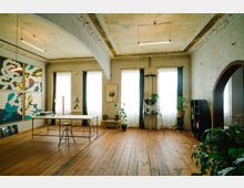 Ein stilvoll eingerichteter Raum mit hohen Decken, Holzboden und großen Fenstern mit weißen Vorhängen, die Tageslicht hereinlassen. Der Raum verfügt über einen Schreibtisch mit einfachen Stühlen, eine Wand mit abstrakten Kunstwerken, Pflanzen und Regale; im Hintergrund ist ein dekorativer Bogen sichtbar.