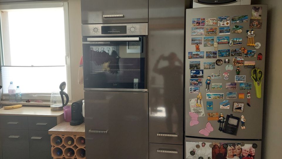 Moderne Küche mit grauen Schränken, Arbeitsplatte und einem Fenster über der Spüle. Rechts stehen ein Hochschrank mit integriertem Backofen sowie ein Kühlschrank, dessen Tür mit vielen Magneten, Postkarten und kleinen Deko-Anhängern bedeckt ist.