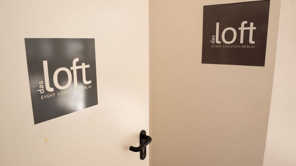 Weiße Innenraumtür und angrenzende Wand mit schwarzen Schildern „das loft – Event Location Berlin“. Die Tür hat eine schwarze Klinke und führt in einen schlichten Flur- oder Eingangsbereich.