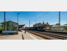 Bahnhof mit mehreren Gleisen und überdachten Bahnsteigen. Ein älteres Gebäude mit graffitibedeckter Fassade und ein verglastes Wartehäuschen befinden sich im Hintergrund, umgeben von elektrischen Fahrleitungen unter klarem Himmel.