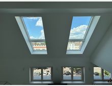 Ein Innenraum mit zwei großen Dachfenstern, die den Blick auf einen blauen Himmel mit einigen Wolken freigeben, sowie drei rechteckige Fenster darunter, durch die eine städtische Straße mit geparkten Autos sichtbar ist. Die Wände sind weiß gestrichen, und das Tageslicht erhellt den Raum.