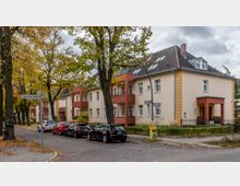 Straßenszene mit mehrstöckigen Wohnhäusern in heller Putzfassade und roten Balkonelementen, umgeben von Bäumen mit Herbstlaub. Parkende Autos säumen die Straße, und Straßenschilder markieren die Kreuzung von Lausanner Straße und Züricher Straße.