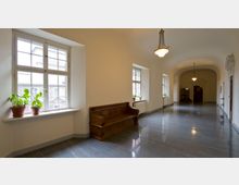 Ein heller Flur in einem historischen Gebäude mit glänzendem Steinboden, Holzbank, Fenstern mit Sprossen und Topfpflanzen auf Fensterbänken. Die Decke ist gewölbt, und hängende Lampen beleuchten den Raum; Türen und Garderoben sind am Ende des Flurs sichtbar.