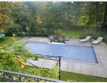 Privater Garten mit einem rechteckigen, abgedeckten Pool, umgeben von einer hölzernen Terrasse. Im Hintergrund befinden sich Bäume, Rasenflächen und zwei Sonnenliegen, die in einer ruhigen, grünen Landschaft angeordnet sind.