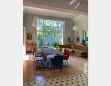 Großes, helles Wohnzimmer mit einer hohen Decke und einer Fensterfront, die auf einen grünen Garten blickt. Der Raum ist mit einem Esstisch, bunten Stühlen, einer Eckcouch, hölzernem Parkettboden und dekorativen Teppichen eingerichtet.