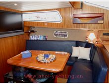 Innenraum einer Yacht mit einer Sitzecke, bestehend aus einer dunkelblauen Polsterbank und einem Holztisch. An der Wand befinden sich ein kleines Fenster, ein Flachbild-TV und eine dekorative Holzskulptur; die Wände und Decken sind mit Holzvertäfelung versehen.