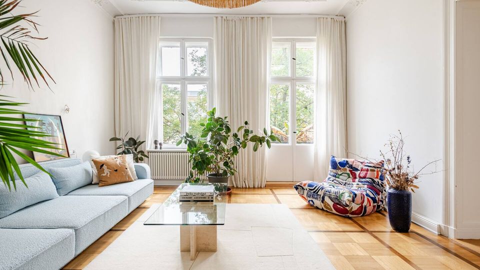 Helles Wohnzimmer mit hohen Decken, Stuckdetails und großem Fenster-/Balkontüren-Ensemble mit weißen Vorhängen. Parkettboden mit hellem Teppich, Glas-Couchtisch, Sofa, bunter Bodensessel sowie mehrere Zimmerpflanzen und eine große runde Deckenleuchte.