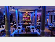 Großes, elegantes Restaurant mit hohen Marmorsäulen, stimmungsvoller blauer Beleuchtung und weiß gedeckten Tischen. Im Hintergrund befindet sich eine zentrale Bar mit dekorativer Beleuchtung und dahinter eine Backsteinwand mit Bogenfenstern.