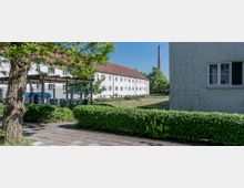 Mehrfamilienhäuser mit weißer Fassade und roten Dächern, umgeben von gepflegten Hecken und einem Rasenbereich; im Hintergrund ist ein hoher Industrieschornstein sichtbar. Links führt ein gepflasterter Gehweg entlang eines Baumes und eines Mülltonnenbereichs.
