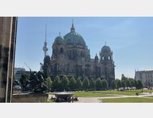 Die Abbildung zeigt den Berliner Dom mit seinen markanten grünen Kuppeln, im Vordergrund eine parkähnliche Rasenfläche mit Bäumen und Spazierwegen. Im Hintergrund ist der Fernsehturm sichtbar, ein bekanntes Wahrzeichen Berlins.