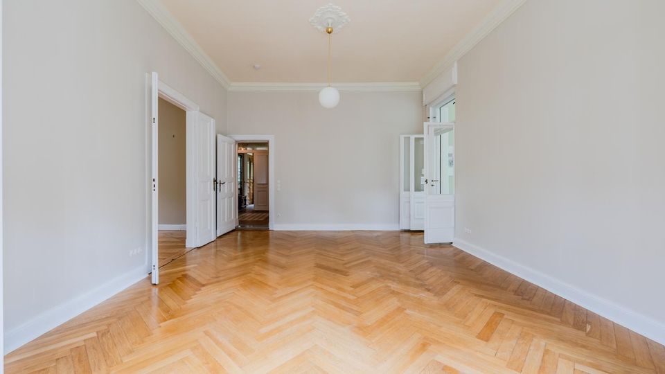 Leerer, heller Wohnraum mit weiß gestrichenen Wänden, Stuckdecke und mittiger Hängeleuchte sowie Fischgrätparkettboden. Mehrere weiße Flügeltüren öffnen zu angrenzenden Räumen und zu einer Fenster-/Balkontür auf der rechten Seite.