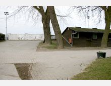 Ein gepflasterter Weg führt an zwei großen Bäumen und einem flachen, dunkelbraunen Holzgebäude mit kleinen Fenstern und Regenrinnen vorbei. Im Hintergrund steht ein weißes Holztor, und am rechten Bildrand ist ein Pflanzenkübel aufgestellt.