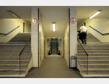 Unterführung mit gelb-weiß gefliesten Wänden, die links und rechts von Treppen mit Metallgeländern sowie in der Mitte von einer Rolltreppe flankiert wird. Ein Deckenlicht beleuchtet den Bereich, und an den Wänden sind Rauchen-verboten-Schilder angebracht.