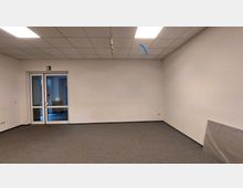 Leerer Raum mit hellen Wänden, grauem Teppichboden und abgehängter Decke mit integrierten Leuchtpaneelen. Im Hintergrund befindet sich eine doppelflügelige Glastür mit Blick in einen weiteren Raum.