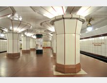 U-Bahn-Station "Altstadt Spandau" mit breiten, weiß verkleideten Säulen, die durch rote Linien akzentuiert sind. Der Boden besteht aus rotbraunen Fliesen, und die Wände sind mit hellen Fliesen mit roten Schriftzügen gestaltet.