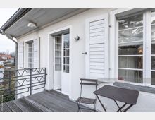 Außenbereich eines Hauses mit heller Fassade, französischen Fenstern und weißen Fensterläden. Der Balkon hat einen Boden aus Holzlatten, ein dunkles Metallgeländer sowie eine einfache Sitzgruppe aus einem Metallstuhl und einem Tisch.