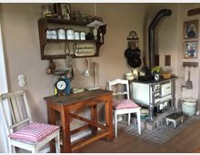 Gemütliche Küche im Landhausstil mit einem alten Holzofen, einem rustikalen Holztisch und zwei Stühlen mit rot-weiß karierten Sitzkissen. An der Wand befinden sich Holzregale mit Vorratsdosen, Küchenutensilien und Wanddekorationen, darunter ein Schild sowie gerahmte Bilder.