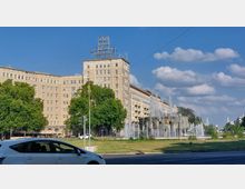 Straßenansicht eines Platzes mit einem großen, historischen Wohngebäude im Hintergrund, gestaltet im Stil des Sozialistischen Klassizismus. Im Vordergrund sind ein Brunnen mit mehreren Wasserfontänen, eine Grünfläche und geparkte Autos zu sehen.