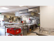 Gewerbliche Küche mit Edelstahl-Arbeitsflächen, professionellen Herdplatten, großen Dunstabzugshauben und Regalen mit Geschirr und Küchenutensilien. Im Hintergrund stehen Kühlgeräte und weitere Aufbewahrungseinheiten, die Wände sind gefliest.