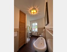 Ein Badezimmer mit Holzfußboden und beigefarbenen Fliesen. Zu sehen sind eine freistehende weiße Badewanne, eine verglaste Duschkabine, ein Waschbecken, eine Toilette und ein Fenster mit halbtransparenter Gardine; die Deckenleuchte hat ein dekoratives Muster.