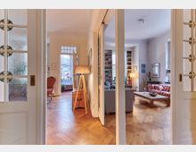 Wohnbereich eines Altbaus mit Holzboden, hohen Decken und großen Fenstern. Im Vordergrund eine geöffnete Tür mit verzierten Glaselementen, dahinter ein Raum mit farbenfrohem Sofa, Bücherregalen und dezenten Holzmöbeln.