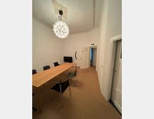 Ein schlichter Konferenzraum mit einem länglichen Holztisch und mehreren schwarzen Stühlen auf einem beigefarbenen Teppichboden. Im Raum befinden sich eine moderne Kronleuchte, eine weiße Tür und ein kleiner Bildschirm.