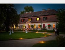 Großes, zweistöckiges Gebäude mit weißer Fassade und rotem Ziegeldach, beleuchtet von warmem Licht aus den Fenstern; vorne ein beleuchteter Garten mit gepflegtem Rasen, dekorativen Kerzen entlang eines geschwungenen Weges und Stehtischen. Im Hintergrund Bäume und im Vordergrund zwei Personen im Gespräch.