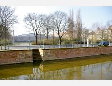 Eine Uferpromenade mit einem kleinen Treppenabgang aus Stein, flankiert von Ziegelmauern, führt zu einem flachen Gewässer. Im Hintergrund sind kahle Bäume, Laternen, eine Straße mit einem geparkten Auto sowie moderne Gebäude sichtbar.