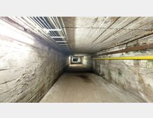 Langer, schmaler unterirdischer Gang mit Betonwänden und -decke, beleuchtet durch Reihen von Leuchtstofflampen. Leitungen und Rohre verlaufen entlang der Decke und Wände, darunter ein gelbes Rohr auf der rechten Seite.