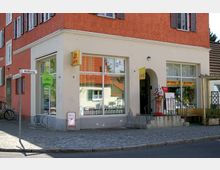 Ecke eines Wohn- und Geschäftshauses mit orangefarbener Fassade, großen Schaufenstern und einem Lotto-Shop im Erdgeschoss. Eine Treppe mit Geländer führt zu einem Eingang, daneben ein Werbeschild in Form eines Eistütenmodells; Straßenschilder und Fahrradständer sind sichtbar.