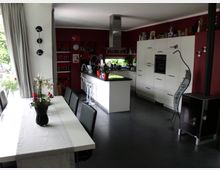 Moderne Wohnküche mit offenem Grundriss, ausgestattet mit weißen Hochglanzschränken, einer zentralen Kochinsel und roten Wandakzenten. Im Vordergrund steht ein Esstisch mit schwarzem Stuhlensemble, daneben ein Holzofen und große Fenster mit hellen Vorhängen, die Tageslicht hereinlassen.