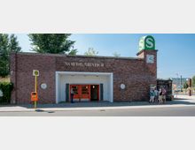 Backsteinbahnhof mit der Aufschrift „Nordbahnhof“, einem großen grünen „S“-Symbol für die S-Bahn auf einem Uhrenturm rechts. Vor dem Gebäude stehen mehrere Personen sowie ein gelber Haltestellenschildpfosten und ein orangefarbener Mülleimer.