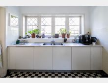 Helle Küche mit einer weißen Arbeitsplatte und einem zentralen Edelstahlspülbecken. Über der Arbeitsfläche befindet sich ein großes Fenster mit sprossenartigem Muster, auf dessen Fensterbank mehrere Topfpflanzen und Dekorationsobjekte stehen; der Boden ist mit schwarz-weiß karierten Fliesen gestaltet.