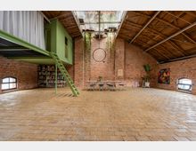 Industrieller Innenraum mit freiliegenden Backsteinwänden, einem hohen Holzdach und einer grünen Podestkonstruktion mit Treppe. Im Raum befinden sich eine lange Tafel mit Stühlen, Pflanzen sowie Regale mit Büchern und Dekorationen.