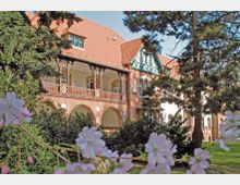 Backstein-Gebäude im Stil der Gründerzeit mit rotem Satteldach, Holzveranda und dekorativen Bögen im Erdgeschoss, umgeben von hohen Bäumen und blühenden Sträuchern im Vordergrund.