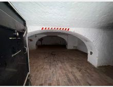 Gewölbekeller mit weiß gestrichenen Ziegeldecken und Ziegelboden, gesehen durch eine geöffnete schwarze Metalltür. Im Hintergrund führt ein weiterer niedriger Rundbogen in einen dunkleren Nebenraum; an der Decke sind rot-weiße Markierungen angebracht.