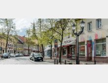 Straßenszene mit gepflastertem Fahrweg, geparkten Autos und Reihen von Geschäften in zweistöckigen Gebäuden mit Schildern und Schaufenstern. Links und rechts der Straße stehen Bäume mit frischem Blätterwuchs und historische Straßenlaternen.