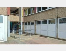 Außenansicht eines mehrstöckigen Betongebäudes mit rauer Fassade und großen, weiß gerahmten Fenster- und Türpaneelen im Erdgeschoss. Vor dem Eingang führt eine Rampe mit Metallgeländer über einen gepflasterten Gehweg; neben dem Durchgang ist eine bunte, mosaikartige Wandfläche zu sehen.