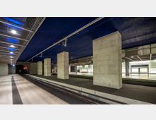 Unterirdischer Bahnhof mit Betonpfeilern, breiten Bahnsteigen und einer blau beleuchteten Decke. Ein Schild mit der Aufschrift „Potsdamer Platz“ ist auf der rechten Seite sichtbar.