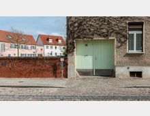 Eine Straßenansicht mit Kopfsteinpflaster, einer Hausfassade mit rauem Putz und einer großen, hellgrünen Holztür, darüber ein Schild mit der Aufschrift „Lindenstraße 10“. Links daneben befindet sich eine rote Backsteinmauer mit rankendem Efeu, dahinter sind Wohnhäuser mit roten Ziegeldächern sichtbar.