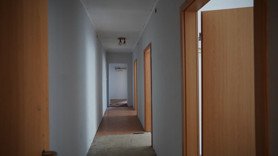 Langer, schmaler Innenflur mit weißen Wänden und mehreren Holztüren auf beiden Seiten. An der Decke hängen einfache Leuchten, der Boden wirkt teils unfertig und am Ende des Gangs ist eine offene Tür mit Hakenleiste sichtbar.