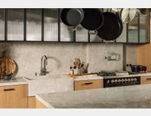 Moderne Küche mit grauer Stein-Arbeitsplatte und Rückwand, hölzernen Unterschränken und mattschwarzen Griffen. Oberhalb hängt ein Gestell mit Pfannen, und ein Gasherd mit goldenen Reglern ist in die Arbeitsfläche integriert.