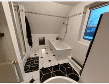 Modernes Badezimmer mit weiß gefliesten Wänden und schräger Decke, ausgestattet mit einer Badewanne, einer Duschkabine und einem Fenster mit Blick nach draußen. Der Boden ist mit schwarz-weißem Fliesenmuster gestaltet, ergänzt durch schwarze Teppiche mit geometrischem Design.