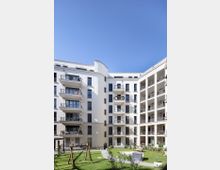 Mehrstöckiges Wohngebäude mit weißer Fassade und Balkonen, die einen begrünten Innenhof mit Gehwegen, Bänken und einem kleinen Spielplatz umgeben. Der Himmel ist klar und blau.