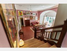 Wohnraum mit rosa Wänden, einer Holztreppe im Vordergrund und einem offenen Bereich, der mit einem Holztisch, Stühlen und einem roten Sofa eingerichtet ist. Der Raum ist mit Kunstwerken, Büchern und Lampen dekoriert und besitzt ein großes Fenster, das viel Tageslicht hereinlässt.