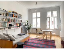 Helles Wohnzimmer mit hohen Decken, Stuck und zwei großen Sprossenfenstern, eines davon geöffnet. Links befinden sich wandmontierte Regale voller Bücher über einem Daybed; im Raum stehen ein runder Tisch mit Hockern, ein Schaukelstuhl, eine Zimmerpflanze und ein gemusterter Teppich auf Holzboden.
